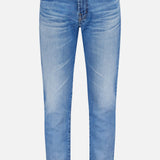 The AG Tellis Aged Jeans are slim-fit, light blue denim with a classic five-pocket design and button closure, shown against a plain white background.