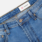 Close-up of Tellis Aged Jeans by AG, featuring blue slim fit denim, waistband, front pockets, metal button, belt loops, and an inner white AG-ED Denim label with a red AG logo.