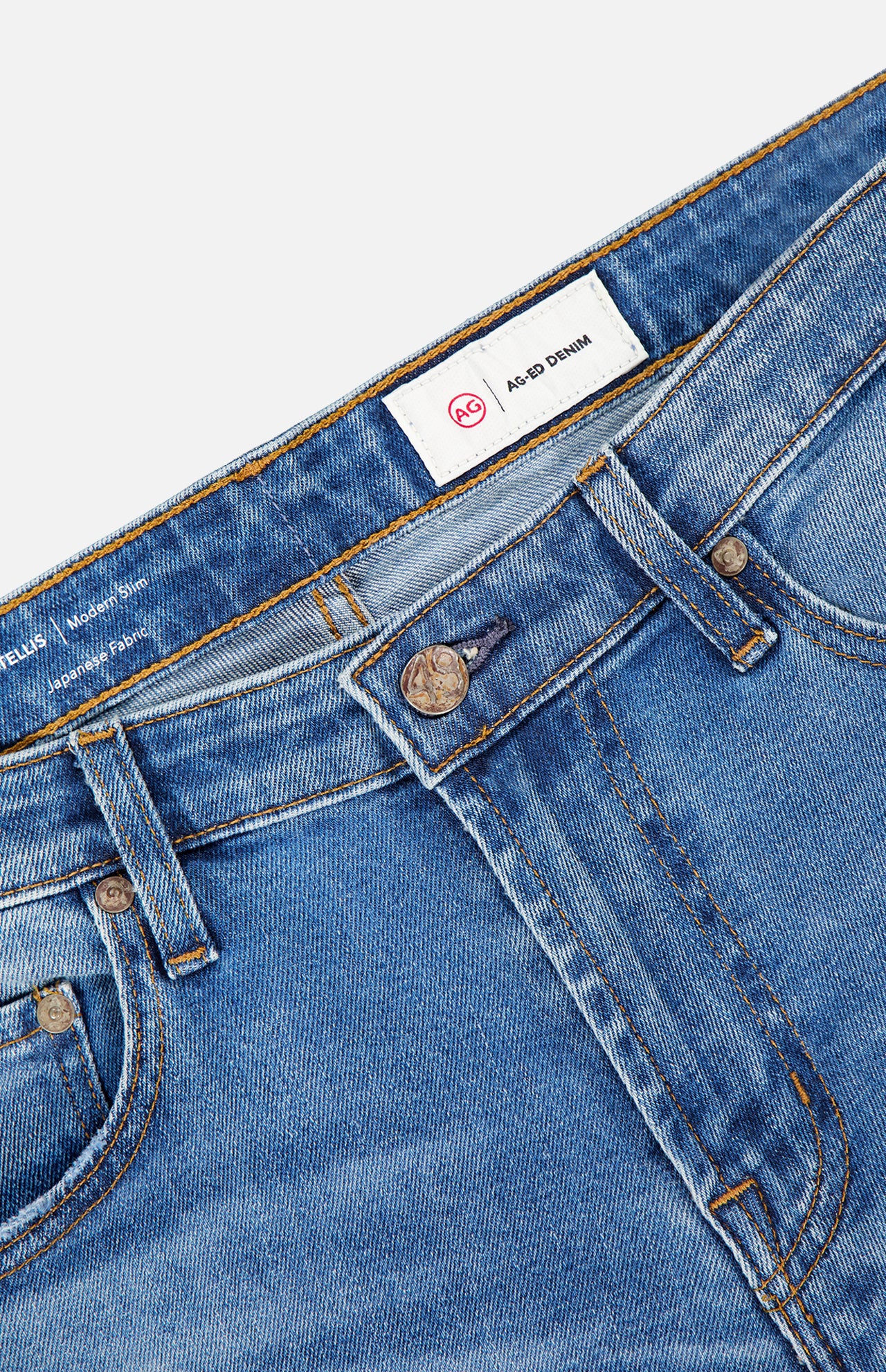 Close-up of Tellis Aged Jeans by AG, featuring blue slim fit denim, waistband, front pockets, metal button, belt loops, and an inner white AG-ED Denim label with a red AG logo.