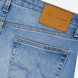 Close-up of the back pocket and waistband of AG Tellis Aged Jeans in slim fit light blue denim, featuring a brown leather patch with branding. Distinctive pocket stitching is shown as the jeans are angled against a white background.