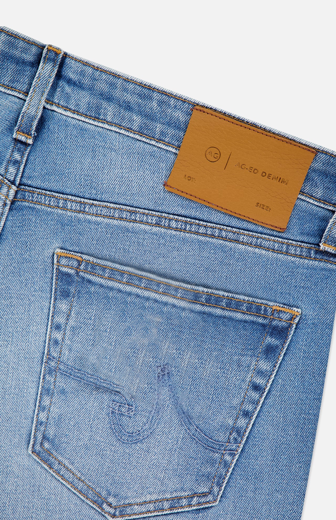 Close-up of the back pocket and waistband of AG Tellis Aged Jeans in slim fit light blue denim, featuring a brown leather patch with branding. Distinctive pocket stitching is shown as the jeans are angled against a white background.