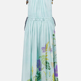 The Alfie Sleeveless Midi Dress by ALEMAIS is a light blue cotton dress with shoulder ties, gathered neckline, flared skirt, floral print featuring colorful leafy accents near the hem, and brown trim along the bottom edge.