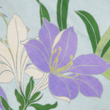 The Alfie Midi Floral Sundress by ALEMAIS features vibrant purple and white flowers with yellow accents and green leaves on a light blue background, capturing the fresh, artistic charm of blooming florals.