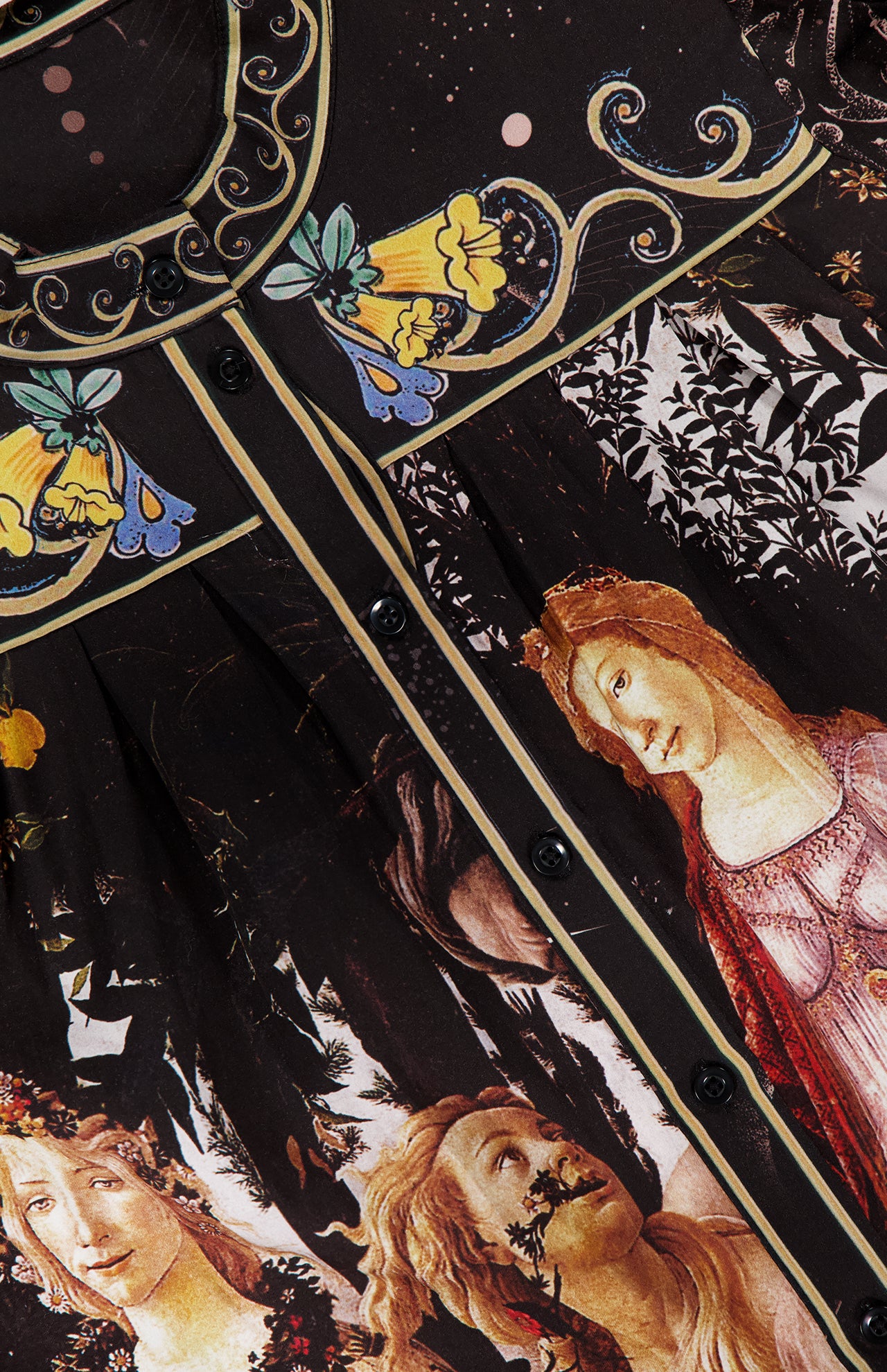 Close-up of the ALICE + OLIVIA Cora Voluminous Long Sleeve Blouse: a black satin top with classical-inspired women’s faces and floral prints in yellow, blue, and green near the collar, blouson sleeves, and black front buttons.