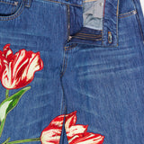 ALICE + OLIVIA’s Low Rise Baggy Jean with Applique in Freyja features a dark blue wash with red and white tulip appliqués on the legs, a relaxed baggy fit, and a partially unzipped waistband that reveals the message “YOU LOOK AMAZING” in red text inside.