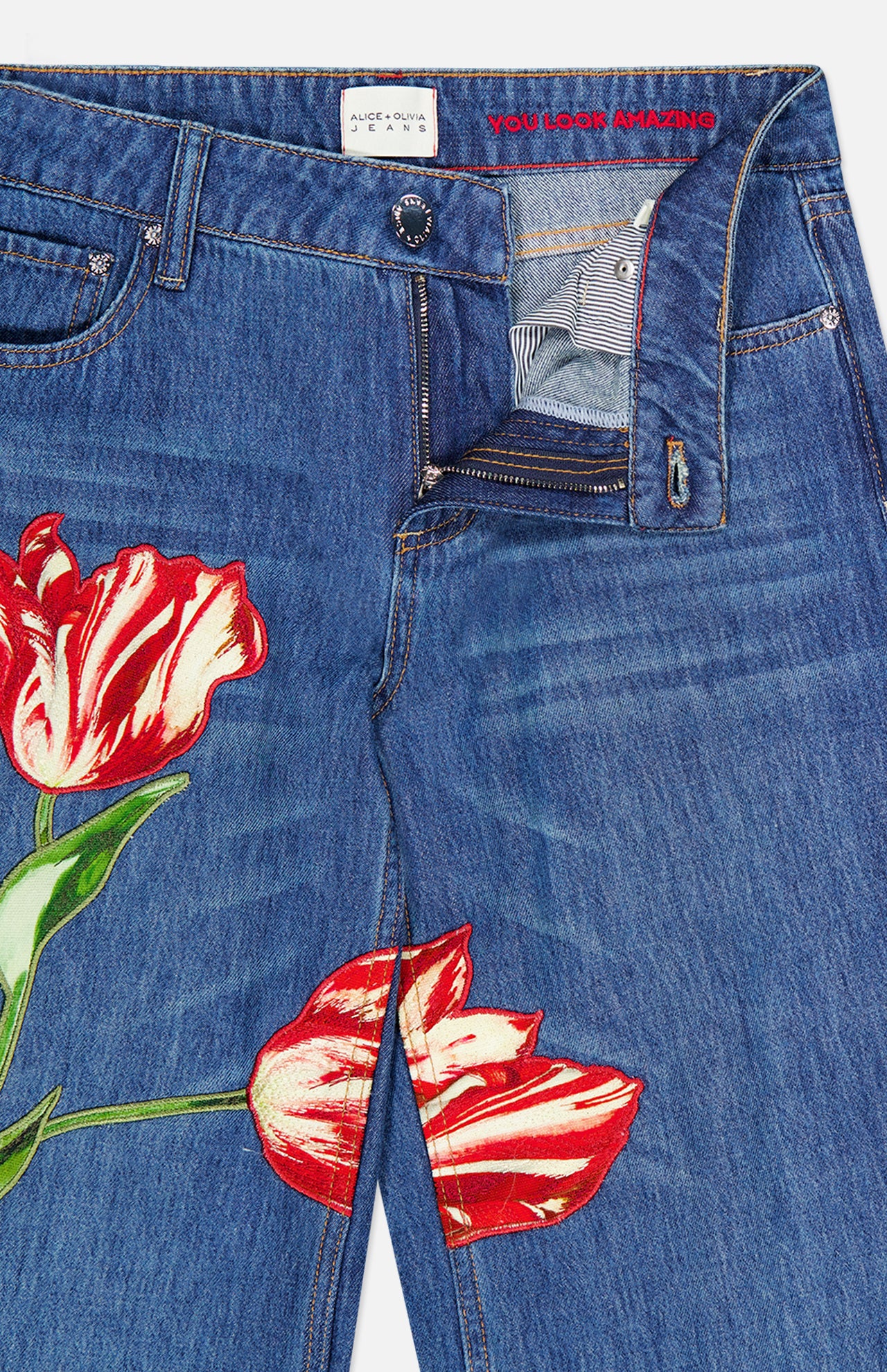 ALICE + OLIVIA’s Low Rise Baggy Jean with Applique in Freyja features a dark blue wash with red and white tulip appliqués on the legs, a relaxed baggy fit, and a partially unzipped waistband that reveals the message “YOU LOOK AMAZING” in red text inside.