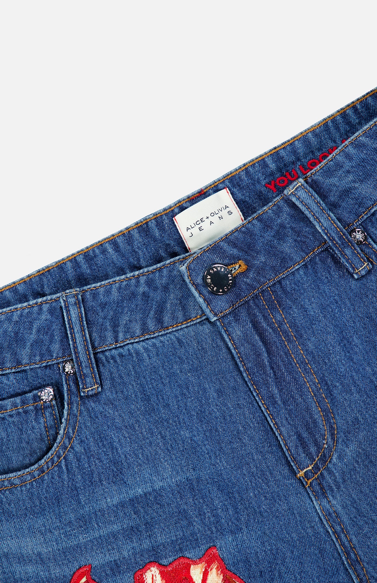 Close-up of the waistband of the Low Rise Baggy Jean with Applique in Freyja by ALICE + OLIVIA, featuring blue dark wash denim, a black button, belt loops, and a white ALICE + OLIVIA JEANS label. Red applique detailing is partly visible on the denim, accentuating the baggy silhouette.