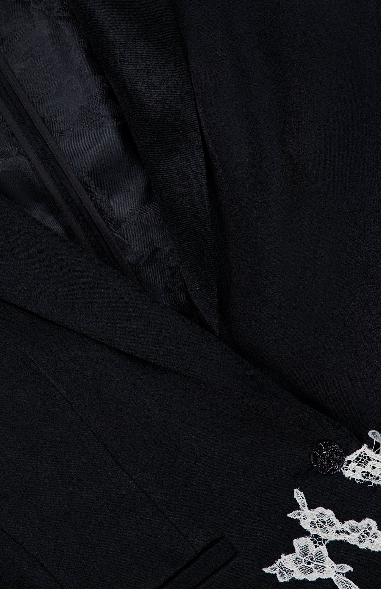 Close-up of the ALICE + OLIVIA Breann Cutaway Lace Long Fitted Blazer in black, featuring decorative cutwork lace trim by the button and a partially visible inner lining accentuating its sculpted silhouette.