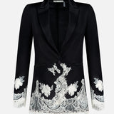 The Breann Cutaway Lace Long Fitted Blazer by ALICE + OLIVIA features a sculpted silhouette with white floral lace trim on the cuffs and hem, single button closure, and peak lapels, shown on a plain white background.
