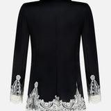 The Breann Cutaway Lace Long Fitted Blazer by ALICE + OLIVIA features black fabric with white cutwork lace trim along the hem and cuffs, creating a sculpted silhouette when viewed from the back.
