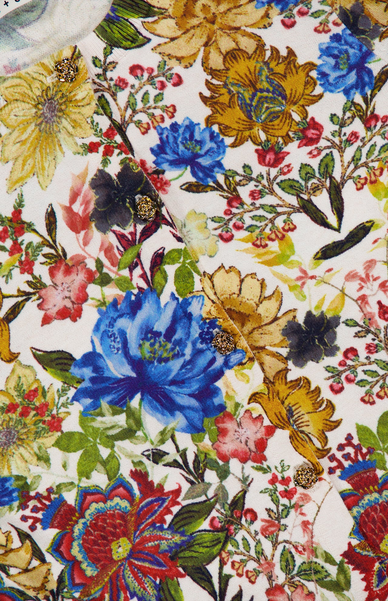Close-up of the Carson Floral Printed Cardigan by ALICE + OLIVIA, showcasing a vibrant floral-patterned fabric with bold blue, yellow, red, and pink blossoms. Sparkling gold buttons are sewn onto the intricate design, adding eye-catching accents—perfect details for this vintage-inspired cropped cardigan that brightens up any fall florals look.