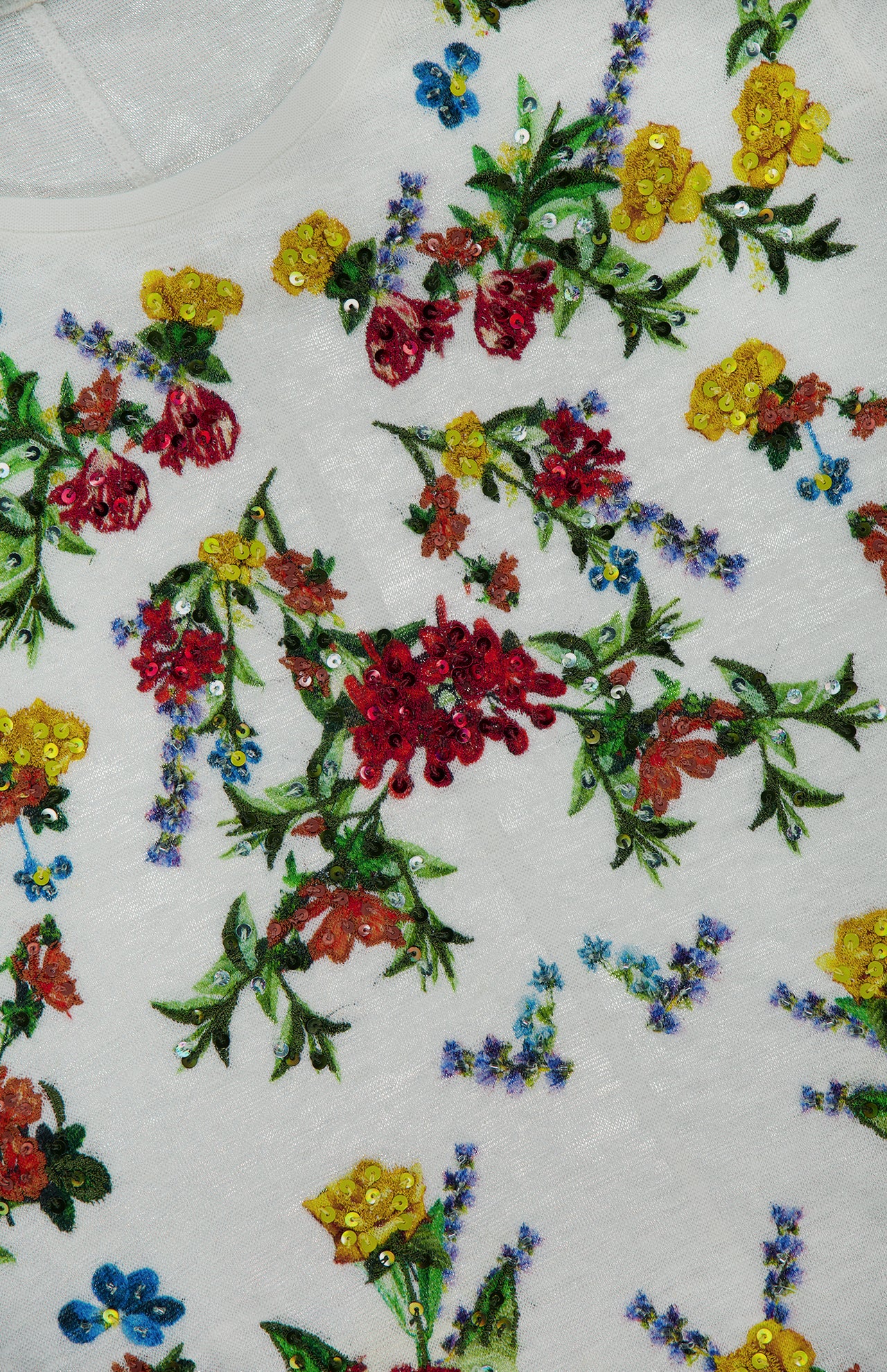 The Cindy Embroidered Cropped T-shirt by ALICE + OLIVIA features white fabric decorated with a vibrant floral design, showcasing red, yellow, orange, and blue flowers with green leaves and scattered sequin embellishments. This cropped t-shirt adds a playful touch to any outfit.