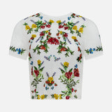 The Cindy Embroidered Cropped T-shirt by ALICE + OLIVIA is a white tee adorned with vibrant floral embroidery in red, yellow, blue, and green across the front, sleeves, and neckline. Delicate floral design details and sparkling sequin embellishments add a touch of shimmer. The t-shirt is displayed against a plain white background.