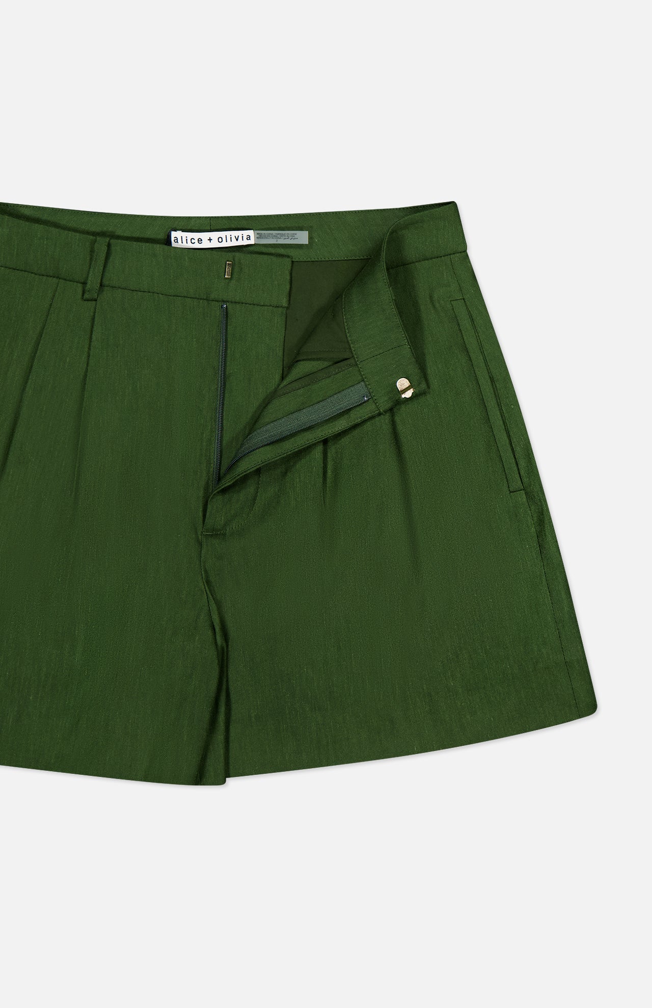 Introducing the Conry Relaxed Long Pleated Short by ALICE + OLIVIA: these dark green linen shorts feature a relaxed fit with front pleats, side pockets, and an open zipper fly. The waistband is finished with a hook-and-bar closure, and the inside tag displays the ALICE + OLIVIA brand name. The shorts are shown on a white background.