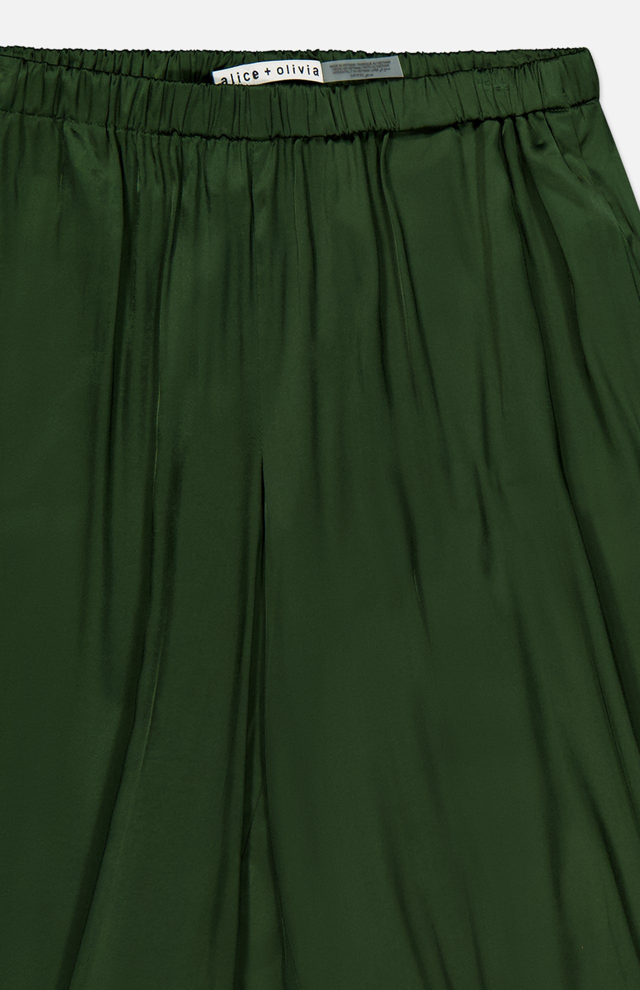 Close-up of the Declan Pull On Genie Pant in dark green silky charmeuse, featuring an elastic waistband and a visible ALICE + OLIVIA label at the top. The shiny fabric has a slightly pleated appearance.