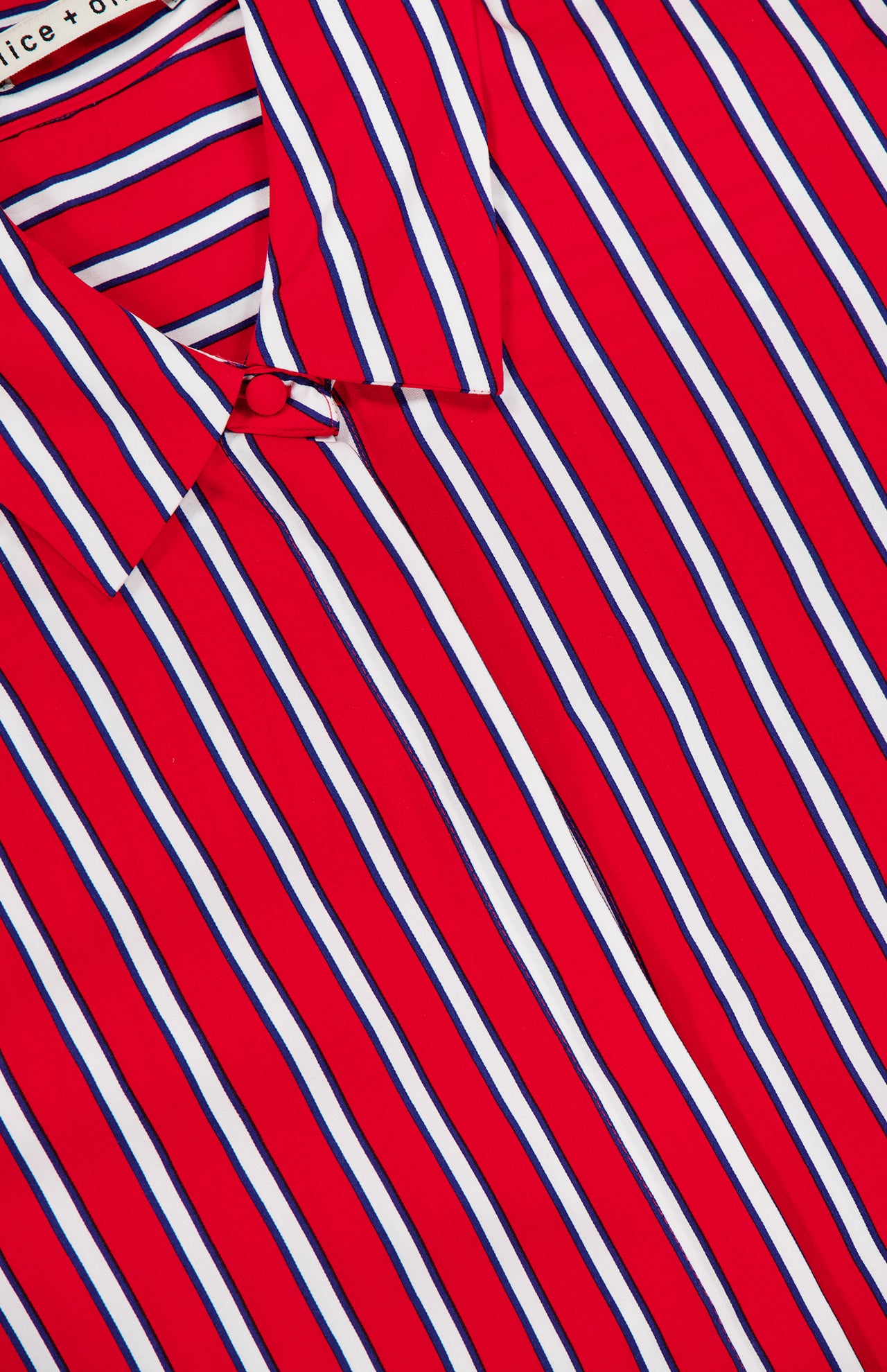 A close-up of the Dimitra Oversized High Low Button Down Shirt by ALICE + OLIVIA in red with vertical white and blue stripes; the top button is fastened, and part of the clothing label appears at the upper left.