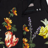 The Dylan High Rise Wide Leg Pant by ALICE + OLIVIA features black linen adorned with a bold dark floral pattern—including yellow roses, purple lavender, orange daisies, and red-striped tulips. The pants are styled partially unzipped to showcase the waistband and button details.