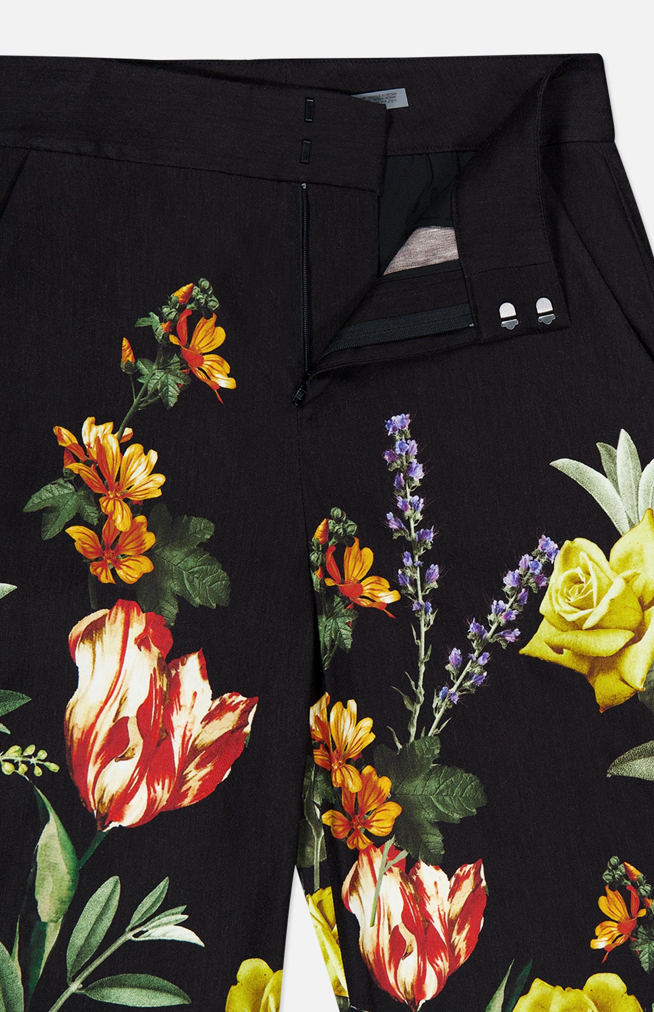 The Dylan High Rise Wide Leg Pant by ALICE + OLIVIA features black linen adorned with a bold dark floral pattern—including yellow roses, purple lavender, orange daisies, and red-striped tulips. The pants are styled partially unzipped to showcase the waistband and button details.