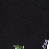 Close-up of the ALICE + OLIVIA Dylan High Rise Wide Leg Pant in black fabric, featuring a single welt pocket and a dark floral pattern with green leaves and purple flowers near the hem.
