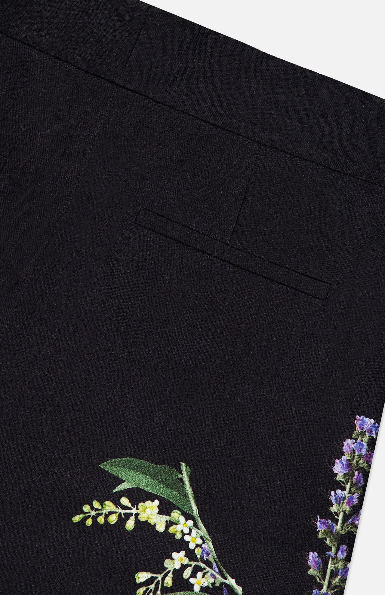 Close-up of the ALICE + OLIVIA Dylan High Rise Wide Leg Pant in black fabric, featuring a single welt pocket and a dark floral pattern with green leaves and purple flowers near the hem.