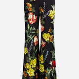 The Dylan High Rise Wide Leg Pant by ALICE + OLIVIA features a black flared silhouette adorned with a vibrant dark floral pattern in red, yellow, and orange flowers with green leaves on both legs, shown against a plain white background.