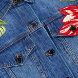 Close-up of the Elijah Embellished Applique Cropped Denim Jacket by ALICE + OLIVIA, showcasing a cropped blue denim silhouette with embroidered red and white tulip floral appliques near the shoulders, metal buttons, and detailed chest pocket stitching.