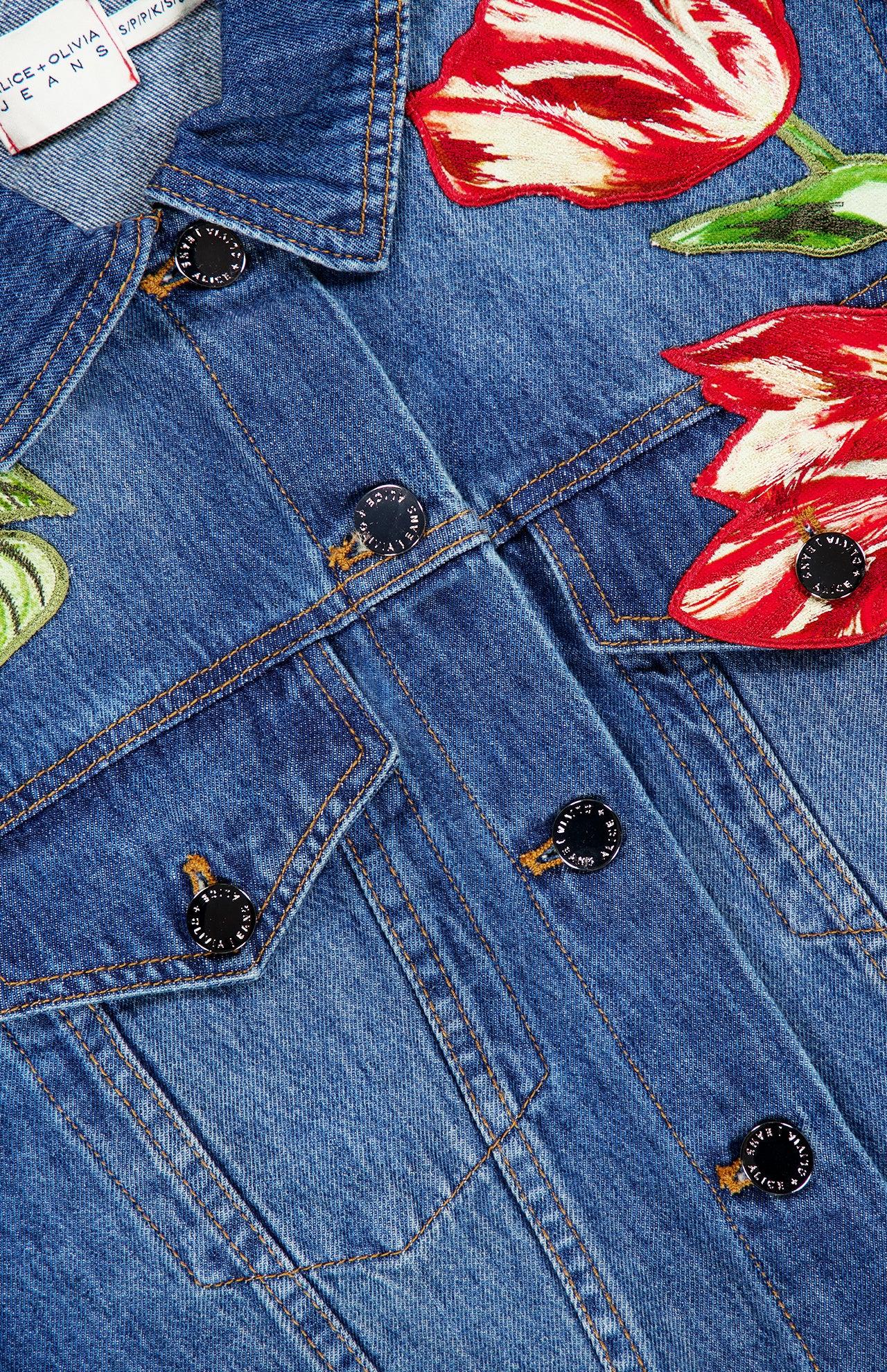 Close-up of the Elijah Embellished Applique Cropped Denim Jacket by ALICE + OLIVIA, showcasing a cropped blue denim silhouette with embroidered red and white tulip floral appliques near the shoulders, metal buttons, and detailed chest pocket stitching.