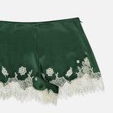 The Elissa Cutaway Lace Short by ALICE + OLIVIA features a dark green satin construction with elegant white bloom cutwork lace trim and detailing along the hem, perfect for channeling a chic summer Hamptons vibe—shown here against a white background.