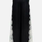 The Olivia Pant with Cutaway Lace Godet by ALICE + OLIVIA features wide-leg black trousers with white cutwork lace on the outer sides, a high waist, and back welt pockets, shown from behind on a white background.