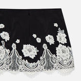 The Rubi Lace Trim Low Rise Micro Skirt by ALICE + OLIVIA features black fabric with white floral lace embroidery, cutwork lace trim, and a scalloped lace hem, shown on a plain white background.