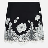 The Rubi Lace Trim Low Rise Micro Skirt by ALICE + OLIVIA is a black micro skirt with white floral lace embroidery, a scalloped fringed hem, and delicate cutwork lace trim for an elegant touch.