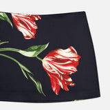 The Rubi Low Rise Floral Skirt by ALICE + OLIVIA is a black mini skirt adorned with a large red and white flower, complete with green stems and leaves printed on the fabric.