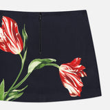 The Rubi Low Rise Floral Skirt by ALICE + OLIVIA features a navy mini silhouette with a black zipper and a striking red and white tulip flower print, accented by green leaves on the front.