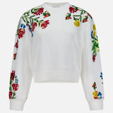 The Russell Embellished Relaxed Crewneck Sweatshirt by ALICE + OLIVIA is a white, cropped-fit sweatshirt adorned with colorful floral embroidery in shades of red, yellow, and blue with green leaves on the shoulders, sleeves, and upper chest.