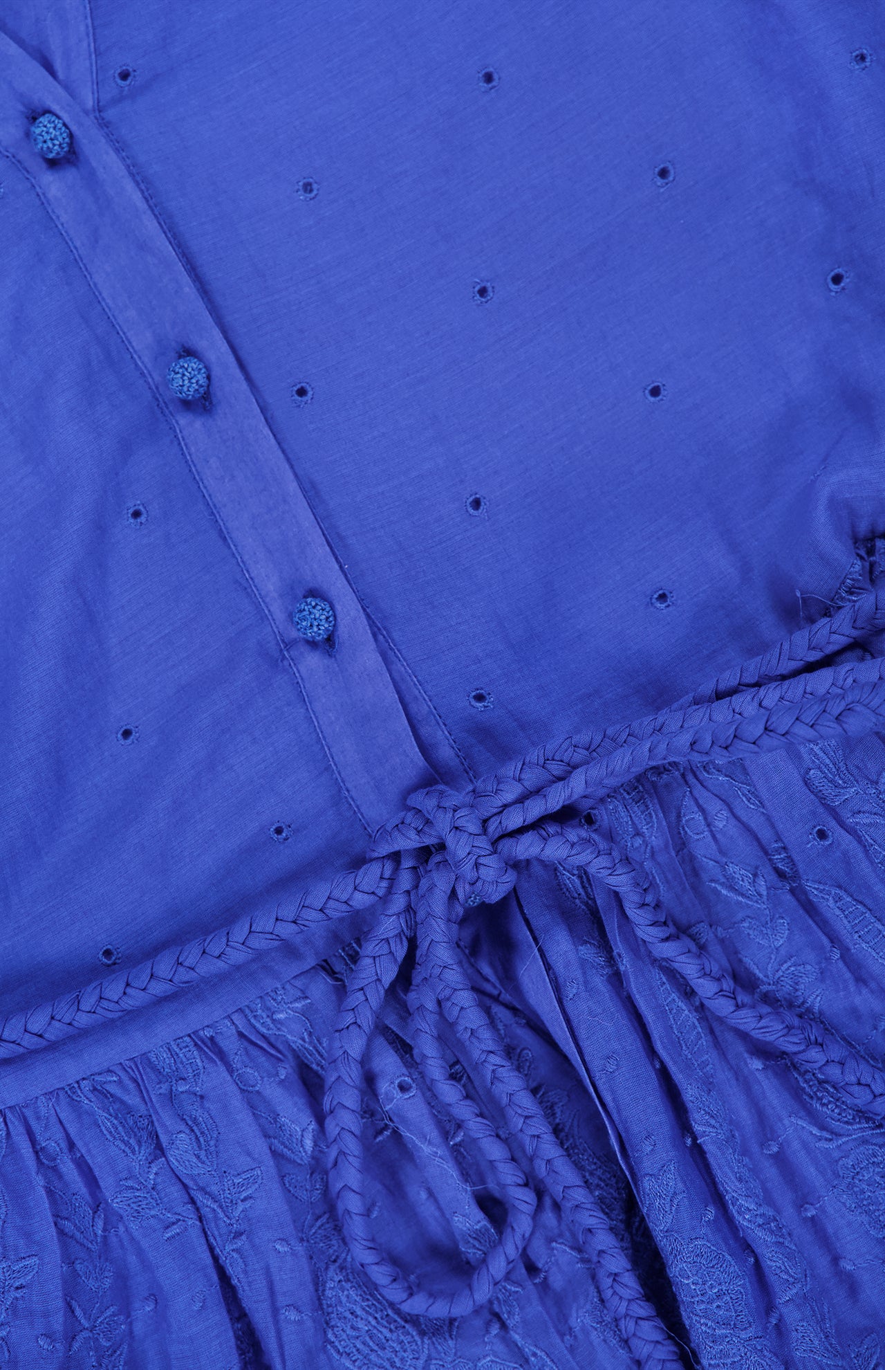 Close-up of the Thalia Puff Sleeve Tunic Dress by ALICE + OLIVIA in cobalt blue, showcasing small embroidered holes in eyelet cotton voile, fabric-covered buttons, a braided waist tie, and a floral lace pattern on the skirt for a bohemian-inspired look.