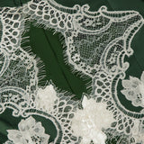 Close-up of delicate white bloom cutwork lace featuring intricate floral and geometric patterns sewn onto the dark green fabric of the ALICE + OLIVIA Thomasina Puff Sleeve Cropped Jacket, highlighting the fine details and texture of both the lace and the smooth material—a romantic touch that adds sophistication to this designer piece.