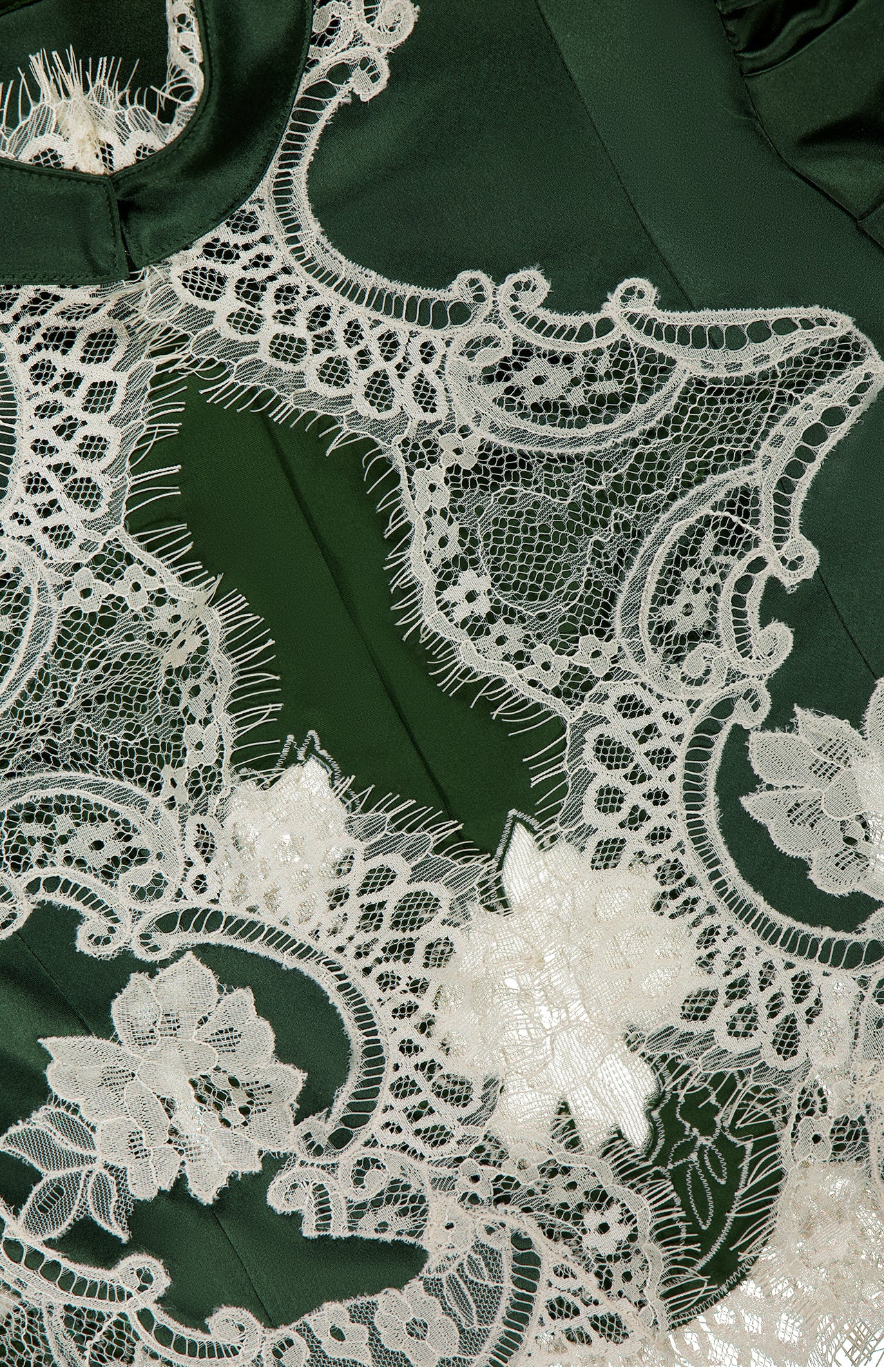 Close-up of delicate white bloom cutwork lace featuring intricate floral and geometric patterns sewn onto the dark green fabric of the ALICE + OLIVIA Thomasina Puff Sleeve Cropped Jacket, highlighting the fine details and texture of both the lace and the smooth material—a romantic touch that adds sophistication to this designer piece.