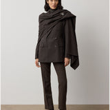 A woman stands against a plain light background, wearing the Mini Herringbone Blazer by RALPH LAUREN COLLECTION—a dark brown wool piece tailored with a draped shawl fastened by a brooch—and paired with leopard print loafers.