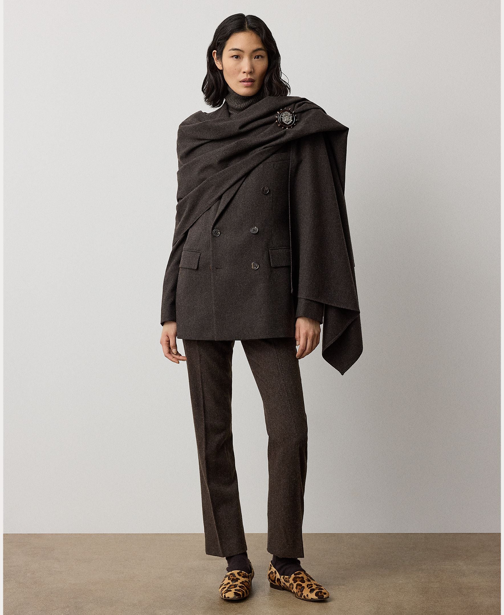 A woman stands against a plain light background, wearing the Mini Herringbone Blazer by RALPH LAUREN COLLECTION—a dark brown wool piece tailored with a draped shawl fastened by a brooch—and paired with leopard print loafers.