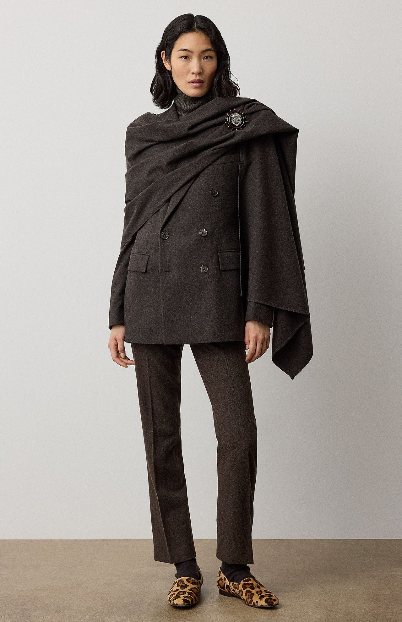 A person wears a dark brown RALPH LAUREN COLLECTION Mini Herringbone Blazer with matching trousers and a detachable scarf draped over their shoulders, secured with a decorative brooch.