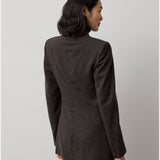 A woman with wavy dark hair, wearing the RALPH LAUREN COLLECTION Mini Herringbone Blazer and matching pants in deep brown, stands facing away from the camera against a plain light background.