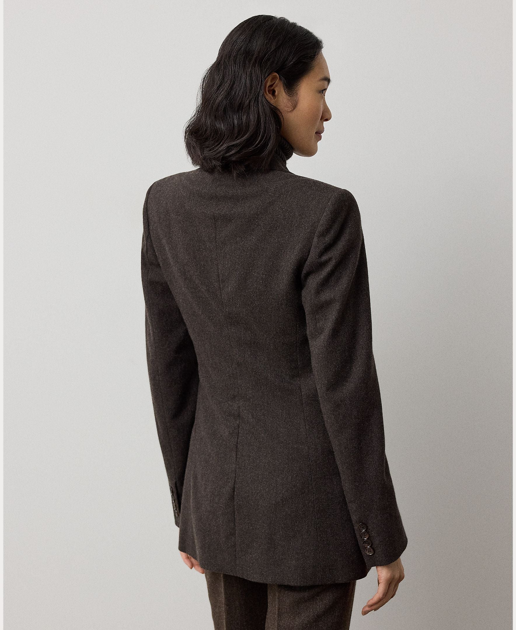 A woman with wavy dark hair, wearing the RALPH LAUREN COLLECTION Mini Herringbone Blazer and matching pants in deep brown, stands facing away from the camera against a plain light background.