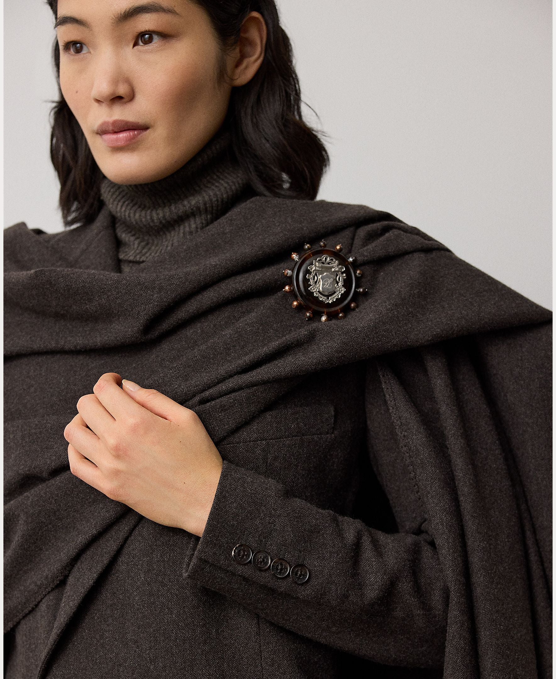 Dressed in a dark turtleneck and the Mini Herringbone Blazer from RALPH LAUREN COLLECTION, accented with a decorative brooch featuring a detailed central emblem and delicate round embellishments, this ensemble exudes refined elegance.