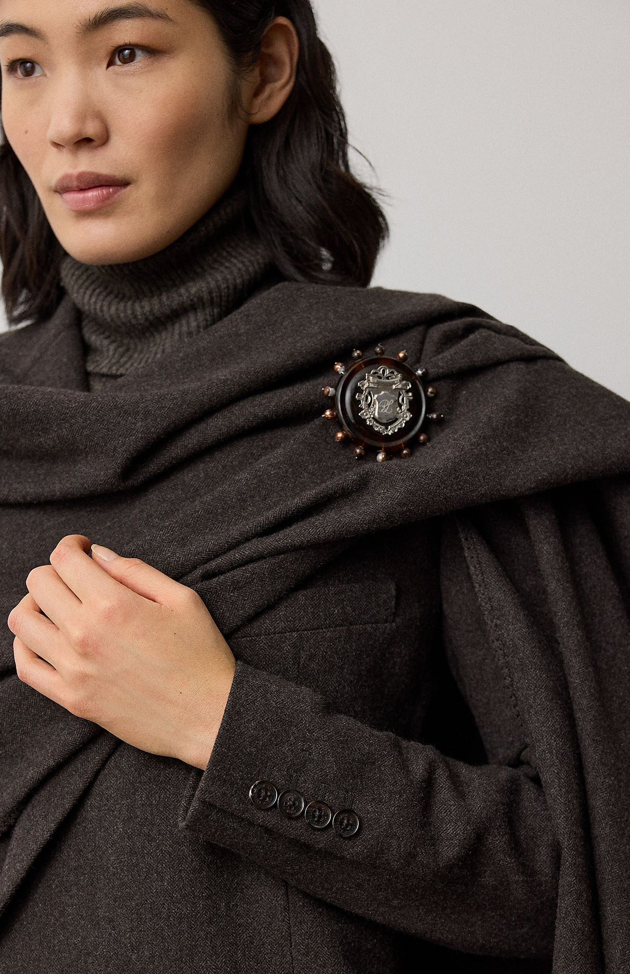 A dark gray RALPH LAUREN COLLECTION Mini Herringbone Blazer worn with a matching detachable scarf pinned with a circular silver and tortoiseshell crest brooch.