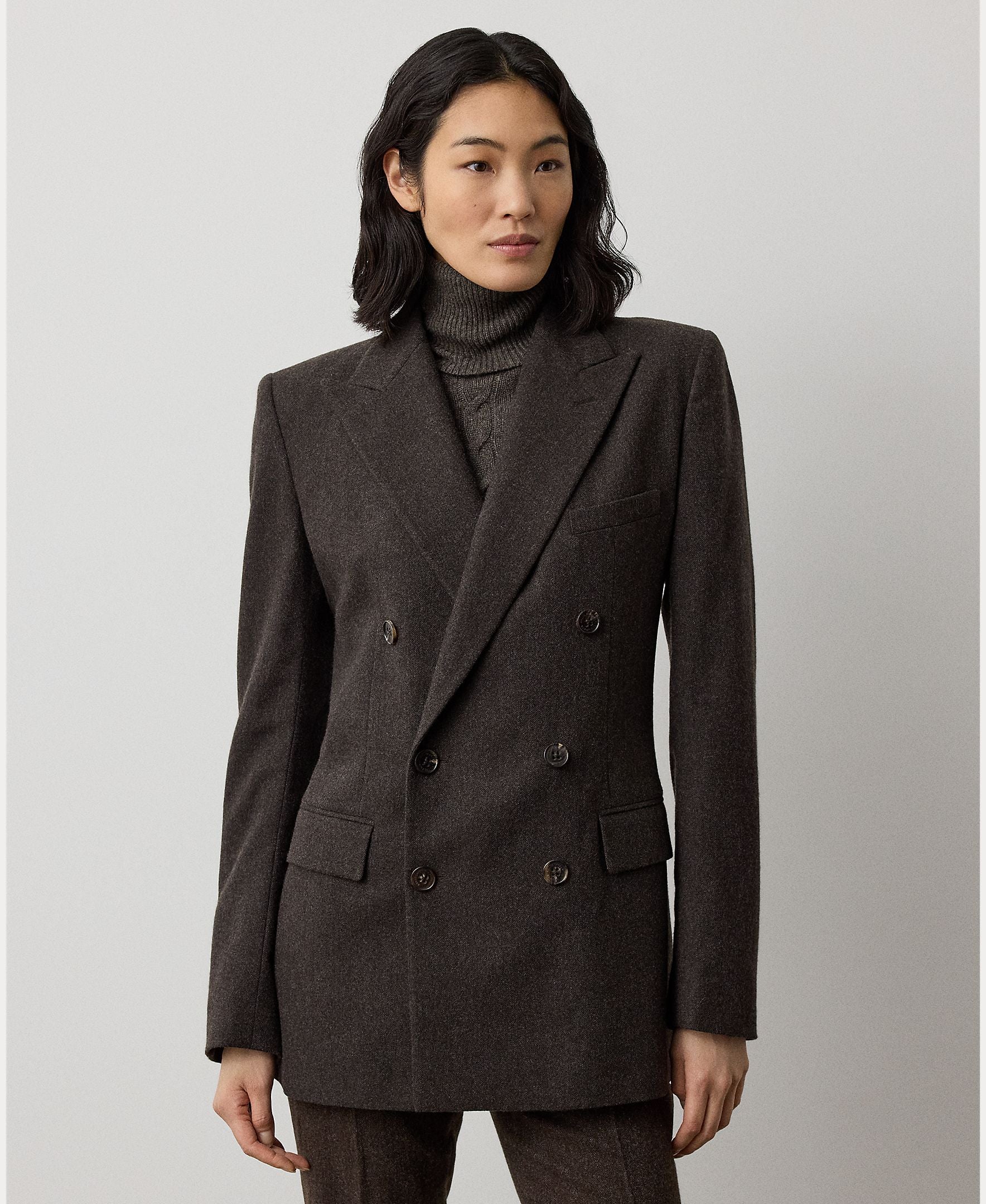 A person with shoulder-length dark hair wears the RALPH LAUREN COLLECTION Mini Herringbone Blazer, featuring a double-breasted design in dark brown wool, styled over a matching turtleneck sweater and standing against a plain light background.