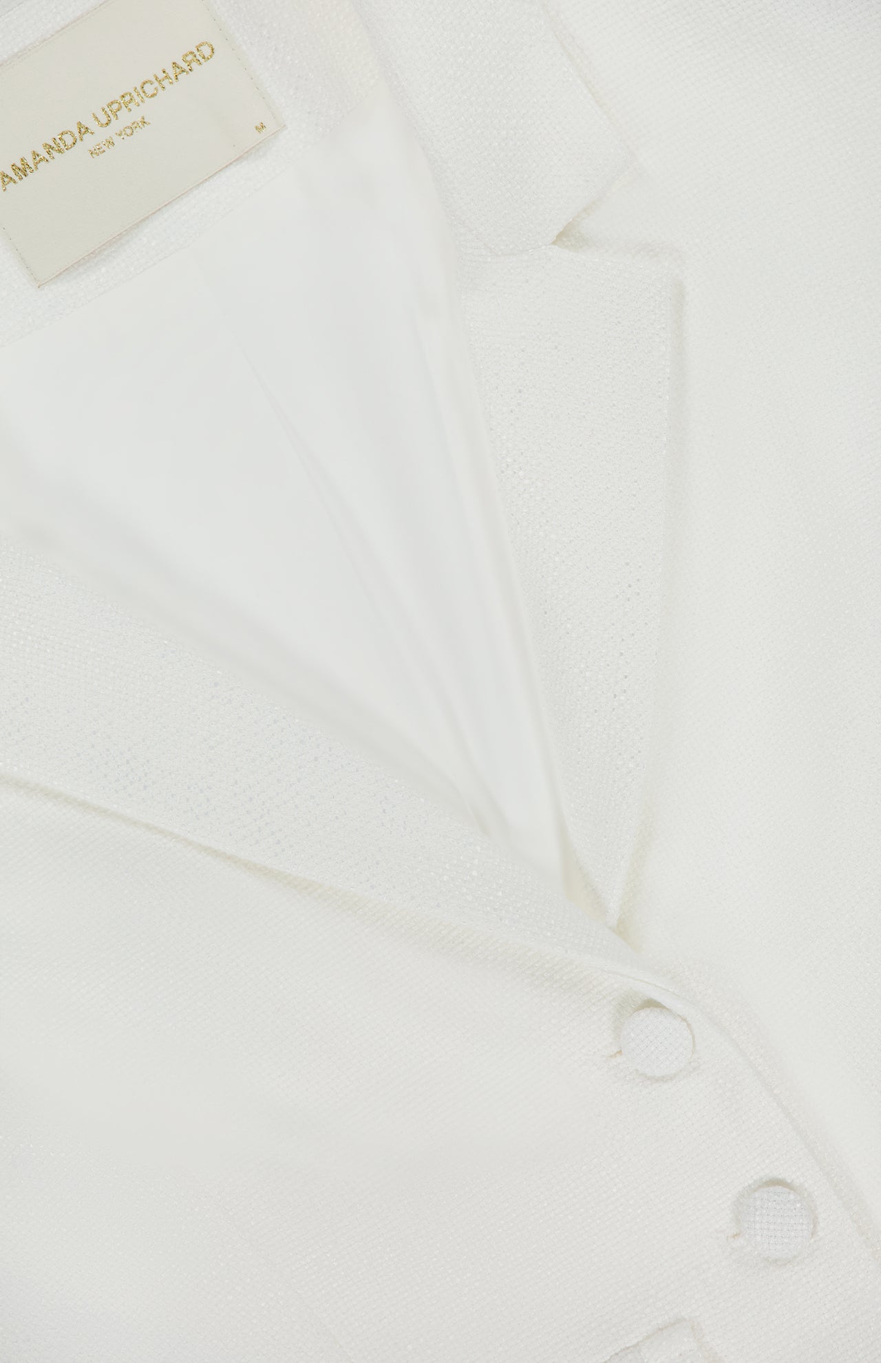Close-up of the Courtney Cropped Jacket by AMANDA UPRICHARD, featuring an ivory hue, notched collar, two matching fabric-covered buttons, and a cropped silhouette. The inner label reads AMANDA UPRICHARD NEW YORK.