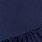 A close-up of the Margo Drop Waist Skirt Dress by AMANDA UPRICHARD shows dark navy fabric with a smooth upper section, a gathered ruffled hem, fine texture, and a subtle sheen.