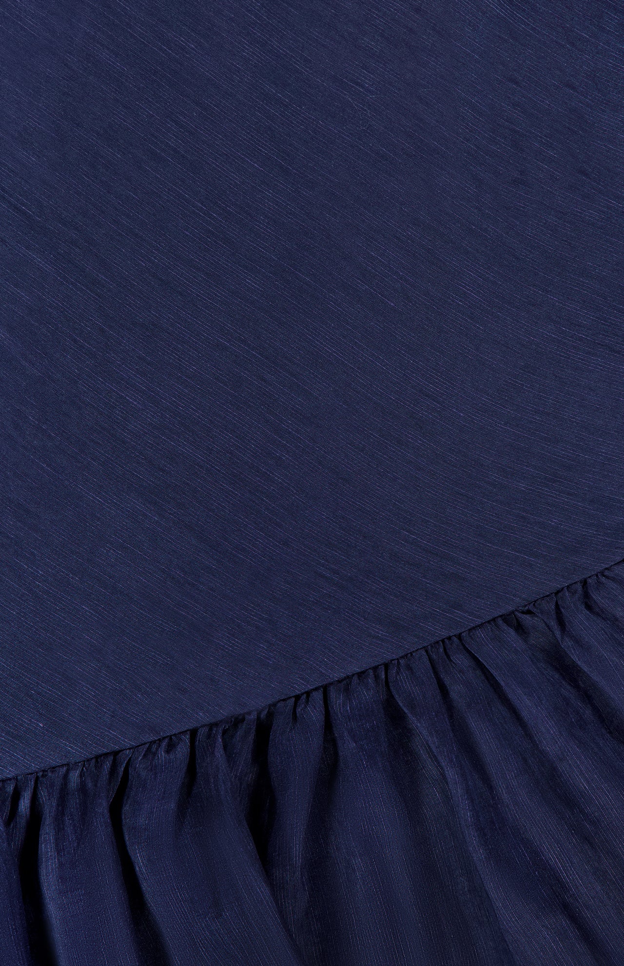 A close-up of the Margo Drop Waist Skirt Dress by AMANDA UPRICHARD shows dark navy fabric with a smooth upper section, a gathered ruffled hem, fine texture, and a subtle sheen.