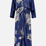 The Olanna Twist Front Maxi Dress by ANDRÉS OTÁLORA is a deep blue satin, long-sleeved dress with a knot front, draped V-neckline, and large light floral print. This Mediterranean-inspired style has a relaxed fit and falls below the knee.