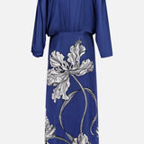 The Olanna Twist Front Maxi Dress by ANDRÉS OTÁLORA features long sleeves, a cinched waist, navy fabric, and white floral patterns on the lower half for a Mediterranean-inspired look.